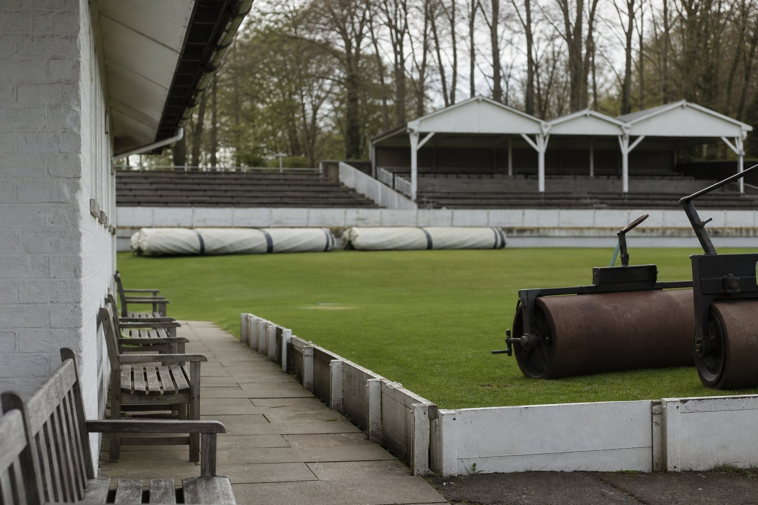 A county cricket ground in flat afternoon light with pitch covers rolled up at the boundary