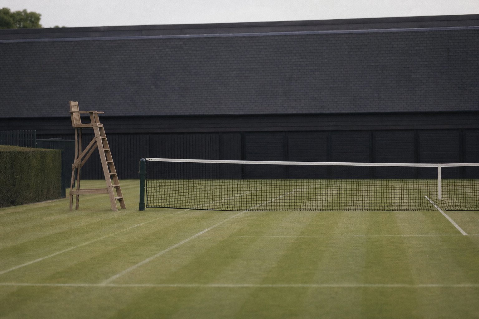 An empty grass tennis court from the side, the net taut and a wooden umpire chair beside it, the lawn clipped in stripes