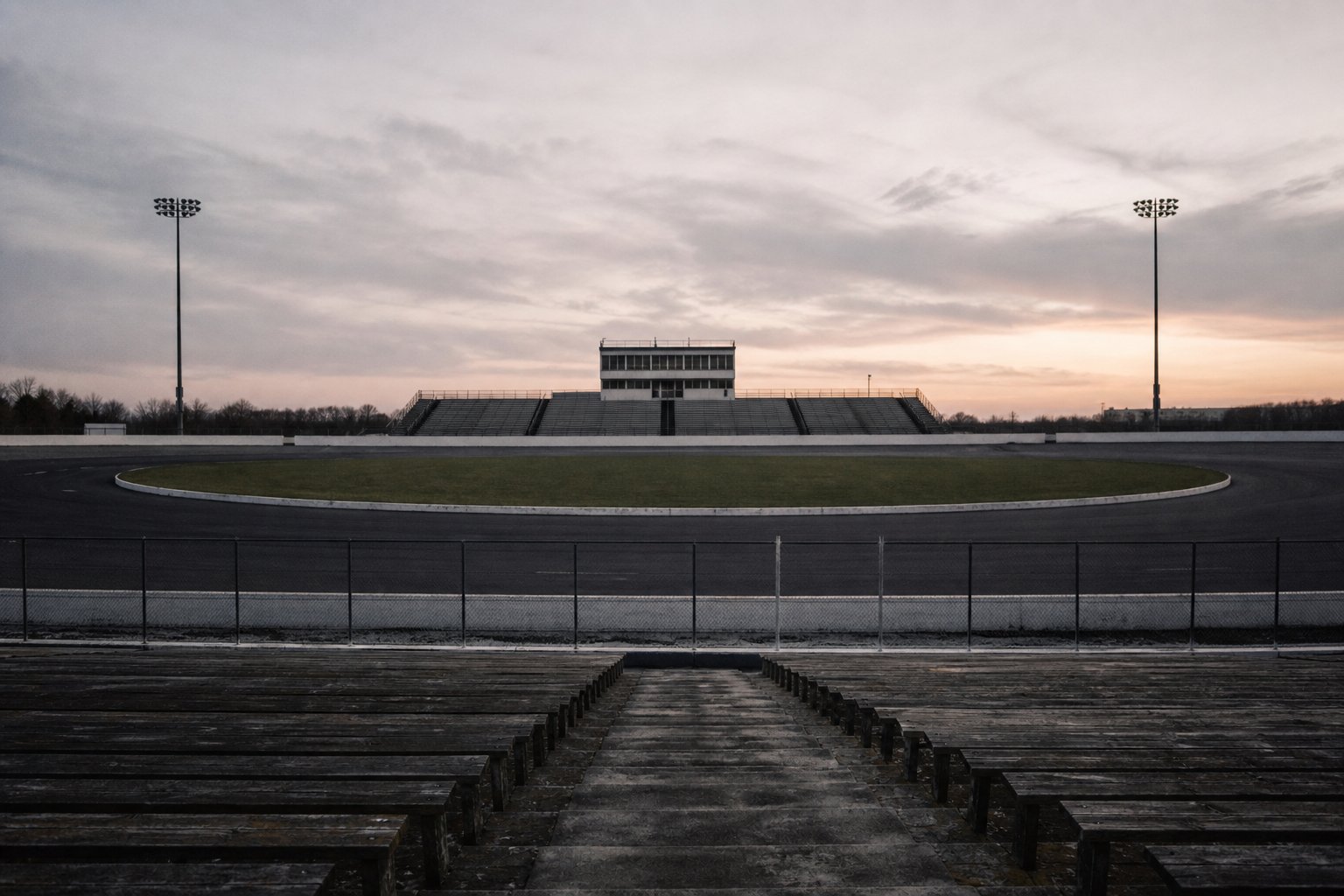 An empty grandstand facing an empty oval speedway at dusk, the timing tower's windows dark