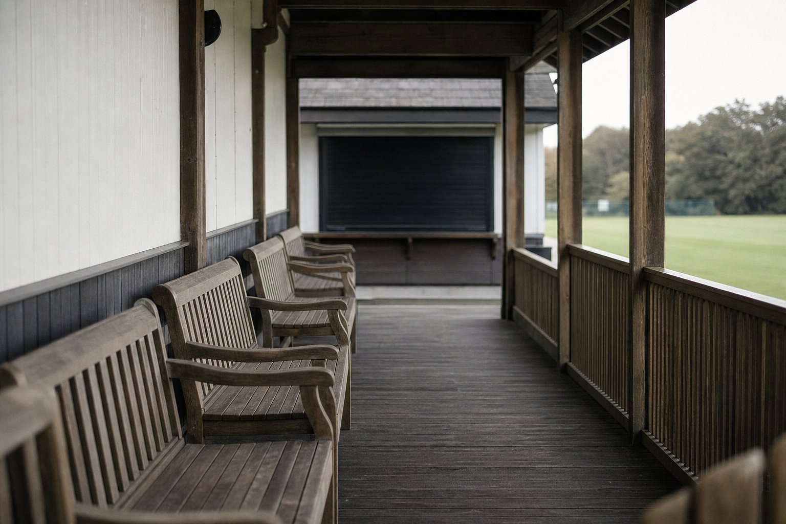 Empty pavilion benches under a wooden verandah at a cricket ground, the scoring hut shuttered at the end